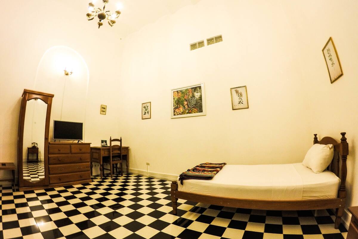 A room with a checkered floor, a bed, a dresser, a chair, a desk, a television, and a chandelier.