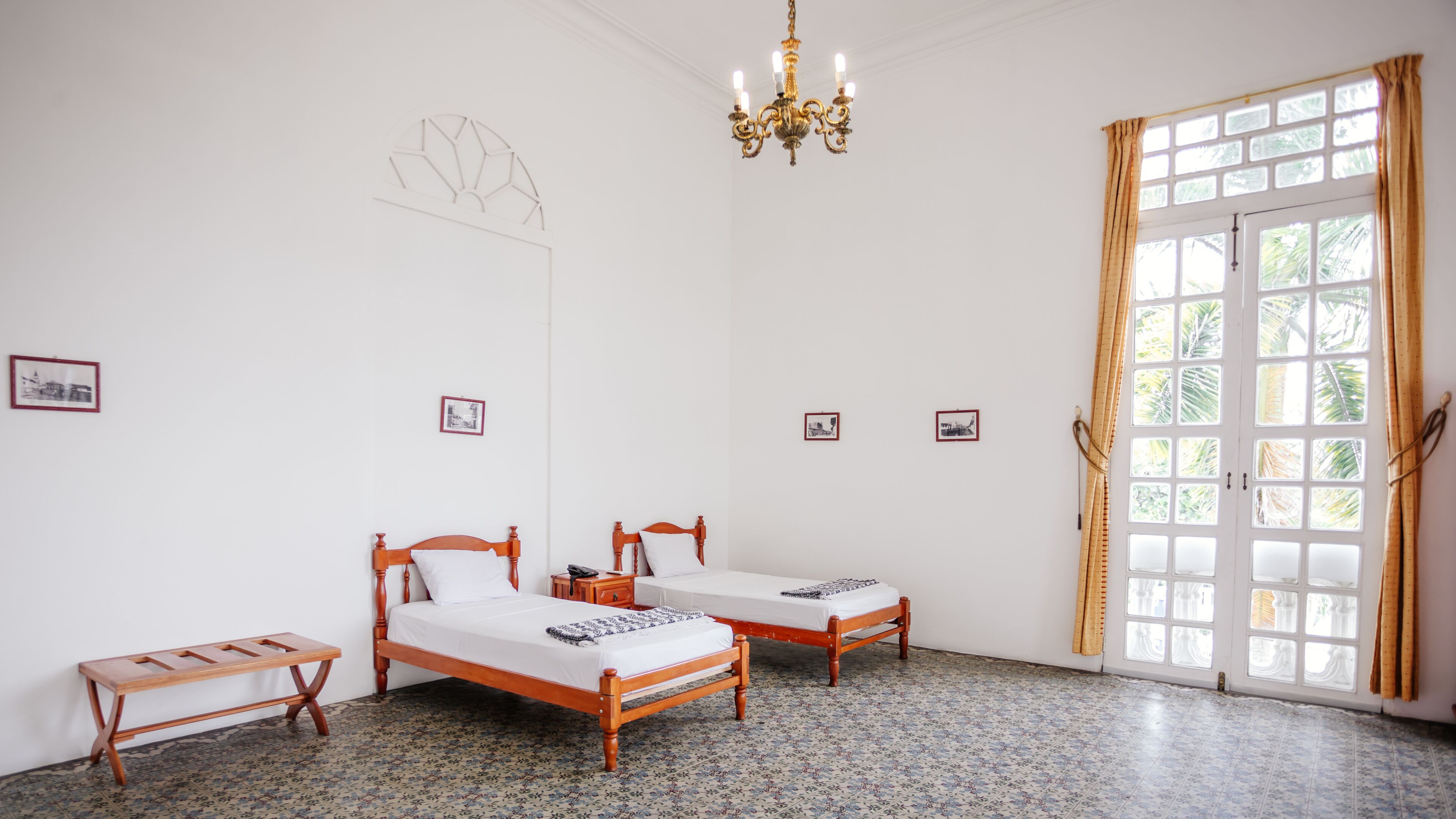 Ein Zimmer mit zwei Holzbett, einer Bank, einer Kristallleuchtre und einem großen Fenster mit Vorhängen.