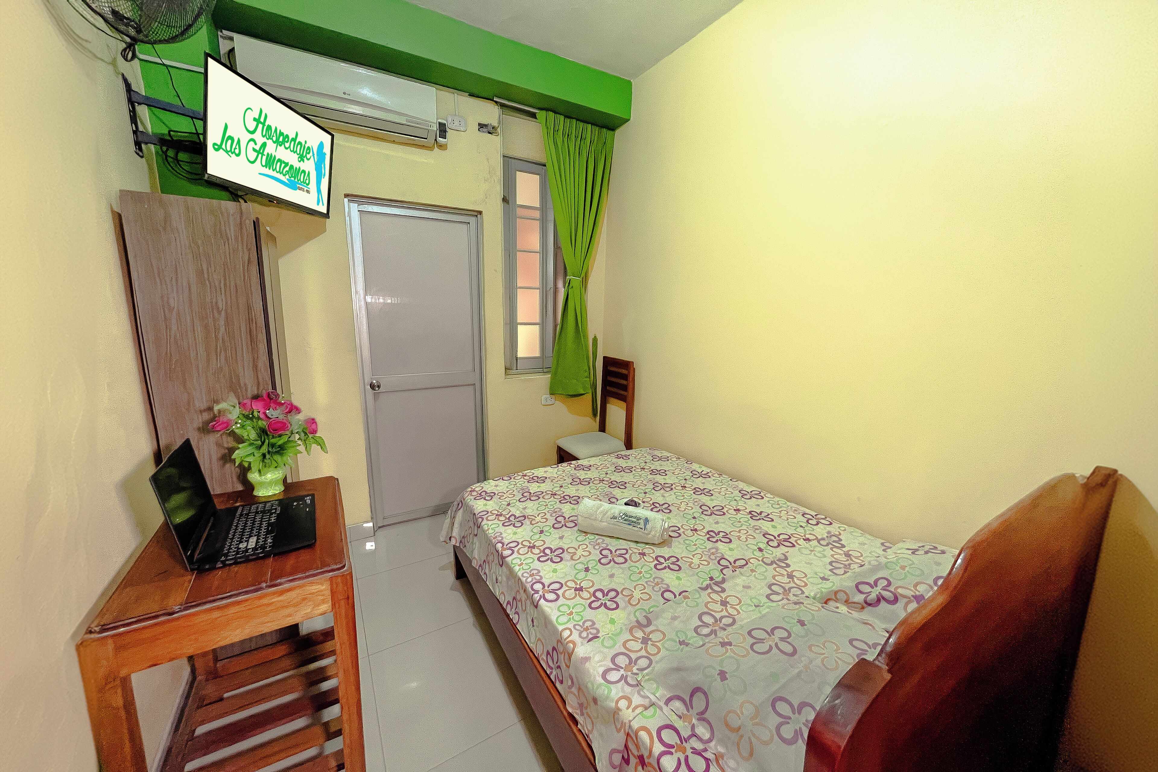 Une chambre d’hôtel avec un lit, un bureau avec un ordinateur portable, un rideau vert et une pancarte au-dessus de la porte.
