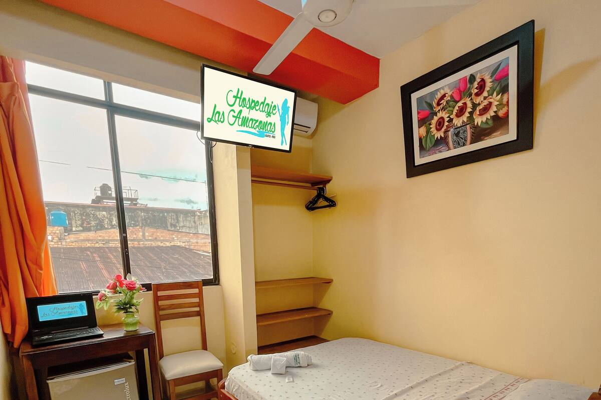 A hotel room with a bed, a desk with a laptop, a chair, a small table with flowers, a framed picture on the wall, and a window with curtains.