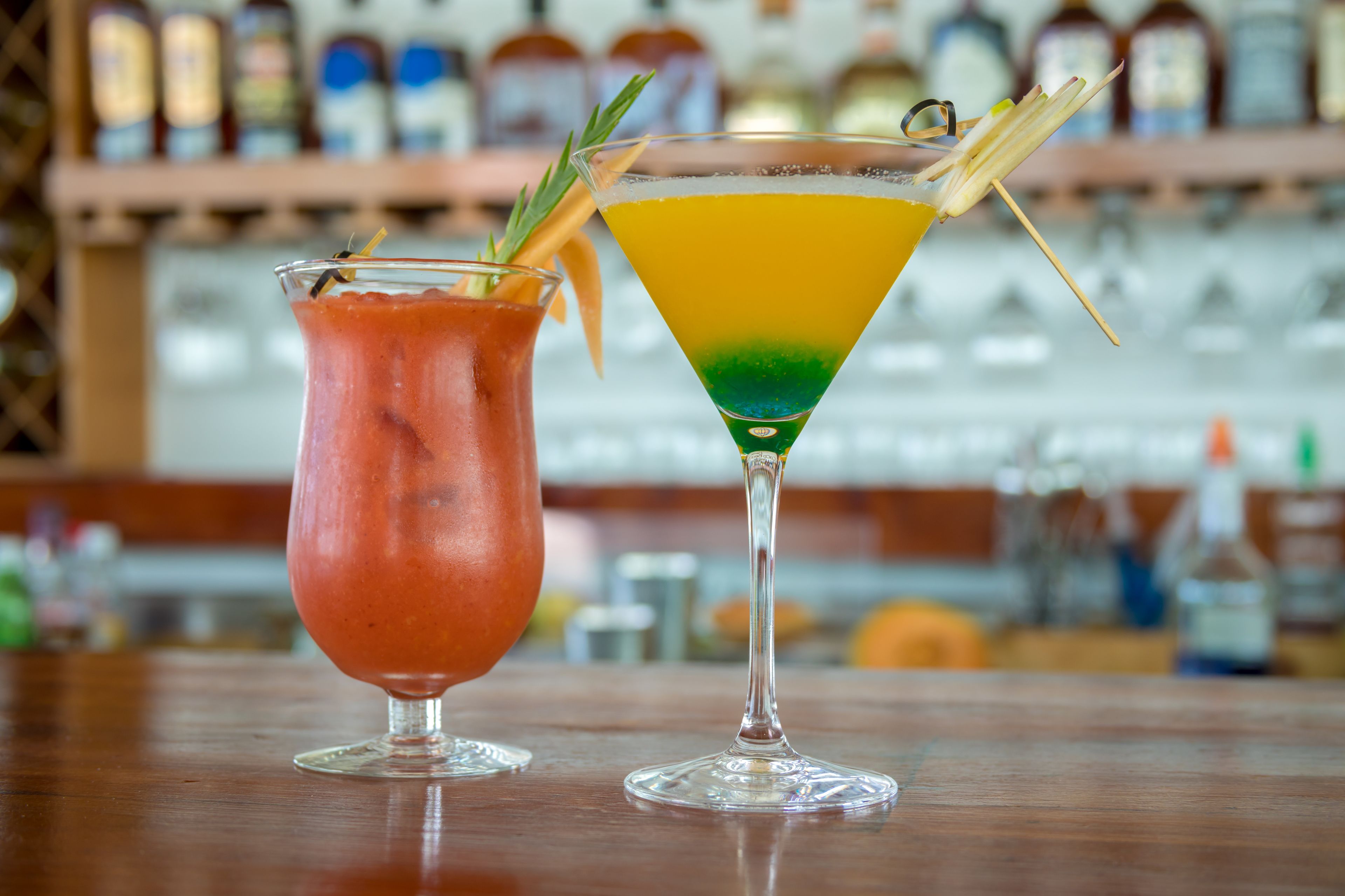 Two cocktails on a bar counter with a blurred background of bottles.