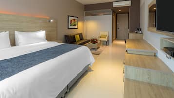 A modern hotel room with a large bed, a sofa, a small table with flowers, a TV, and a desk with a computer.