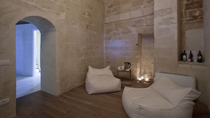 Une pièce aux murs de pierre, avec un plancher en bois, deux fauteuils en forme de sac de haricots blancs, une petite table avec une bouilloire et une étagère remplie de bouteilles.