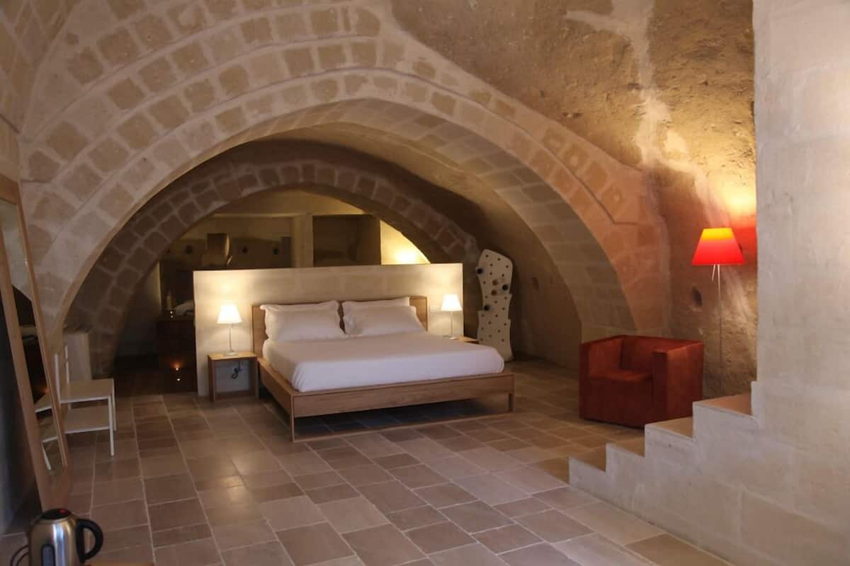 Een slaapkamer met een stenen boog, een bed, een nachtkastje, een lamp, een stoel en een trap.