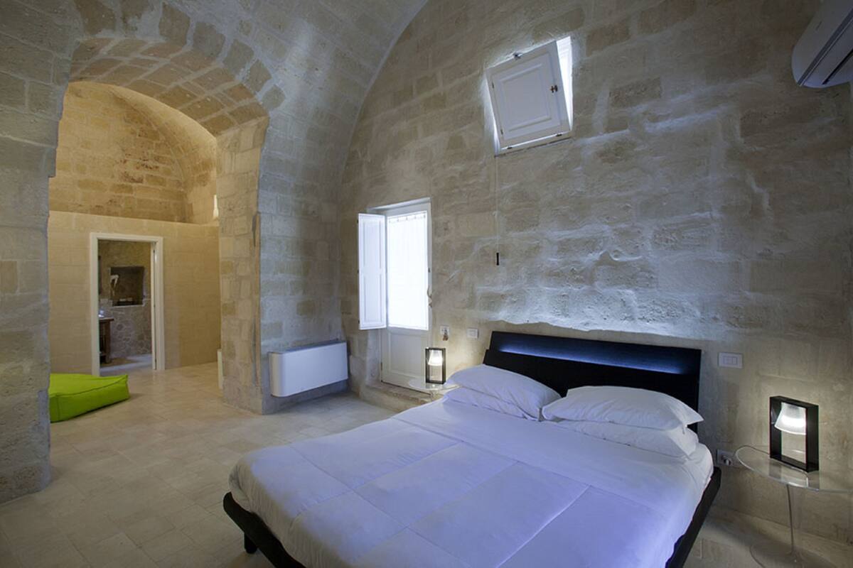 Een slaapkamer met een stenen muur, een bed, een raam en een deur naar een badkamer.