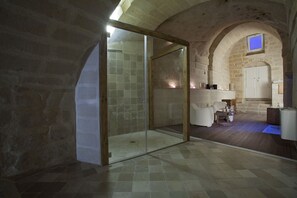 Un baño con paredes de piedra, cabina de ducha con mampara de vidrio, suelo de madera y zona de lavamanos blanca.
