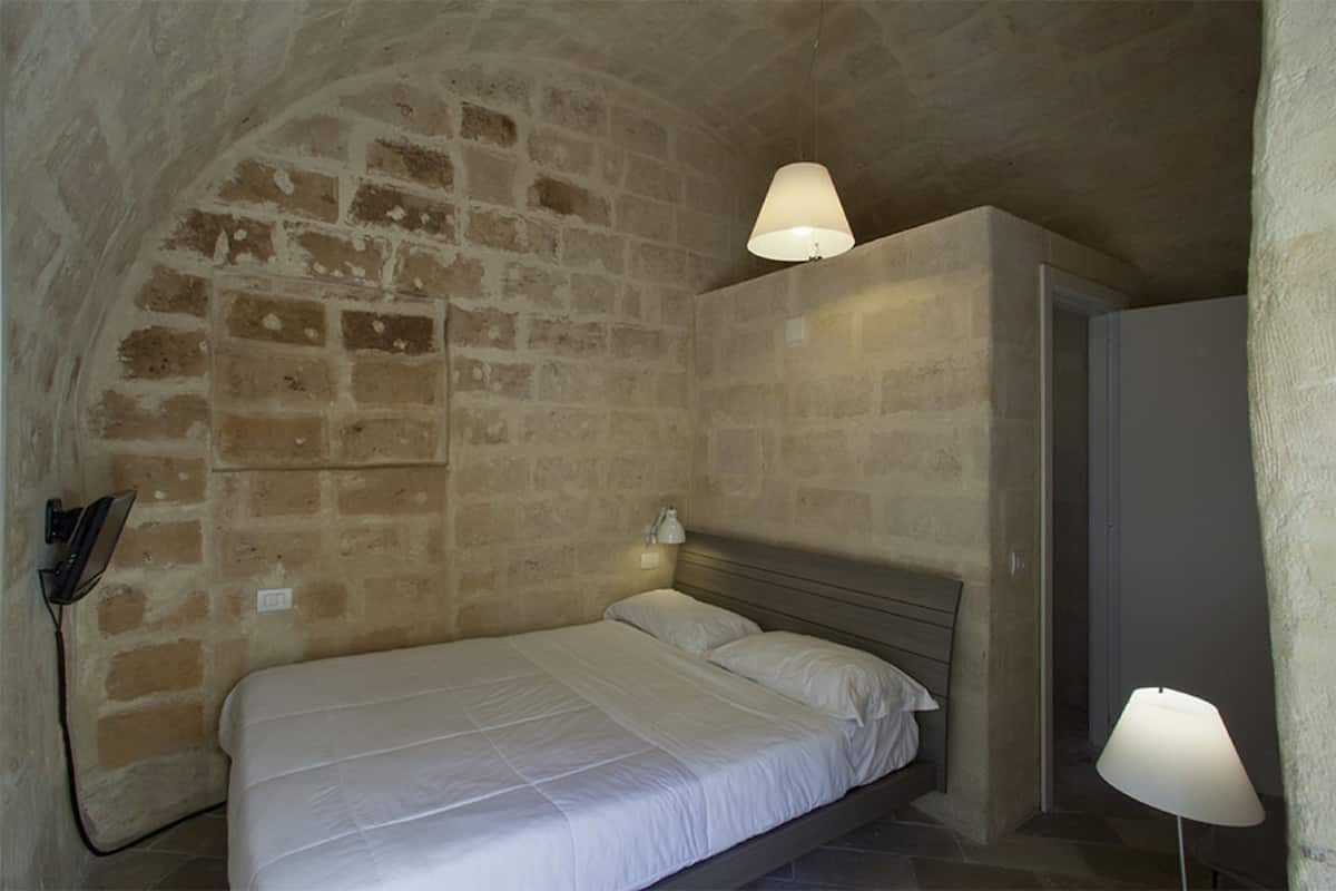 Een slaapkamer met een bakstenen muur, een bed met witte beddengoed, een bedlampje en een hanglamp.