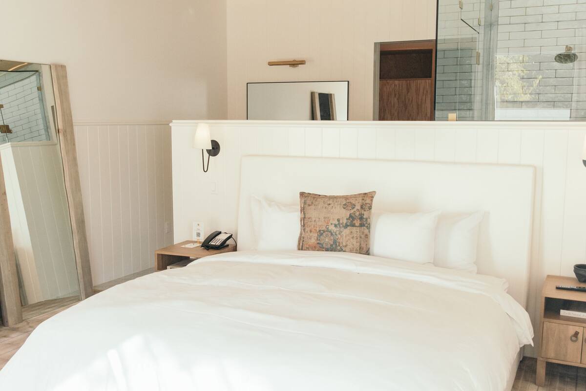 A bedroom with a large bed, a wooden ceiling, a bedside table with a lamp, and a mirror on the wall.