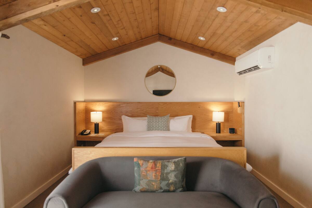 A bedroom with a wooden ceiling, a large bed with white linens, two bedside tables with lamps, and a chair with a cushion.