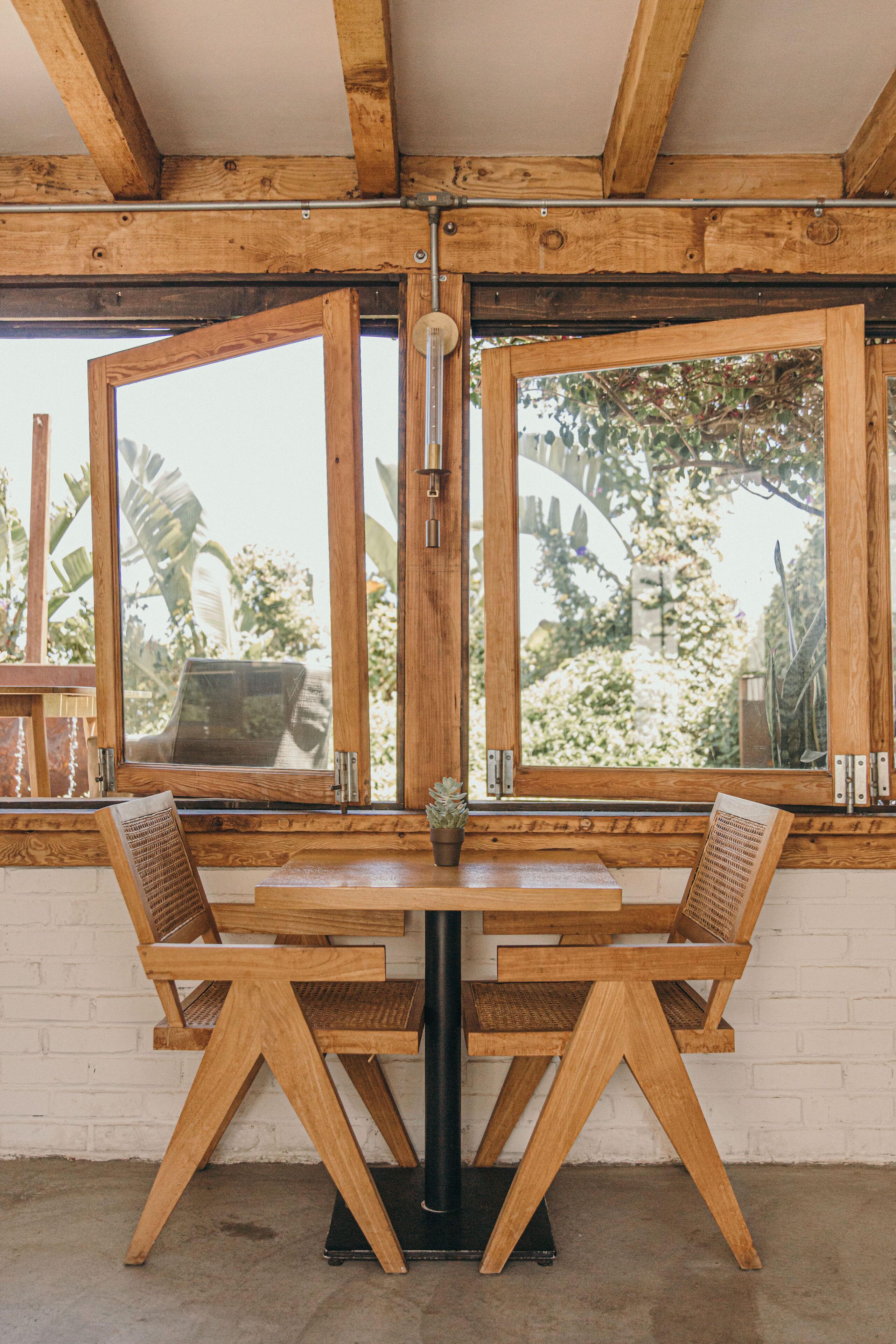 Une table à manger en bois, accompagnée de deux chaises, d’une plante en pot et offrant une vue sur la verdure grâce à de grandes fenêtres.