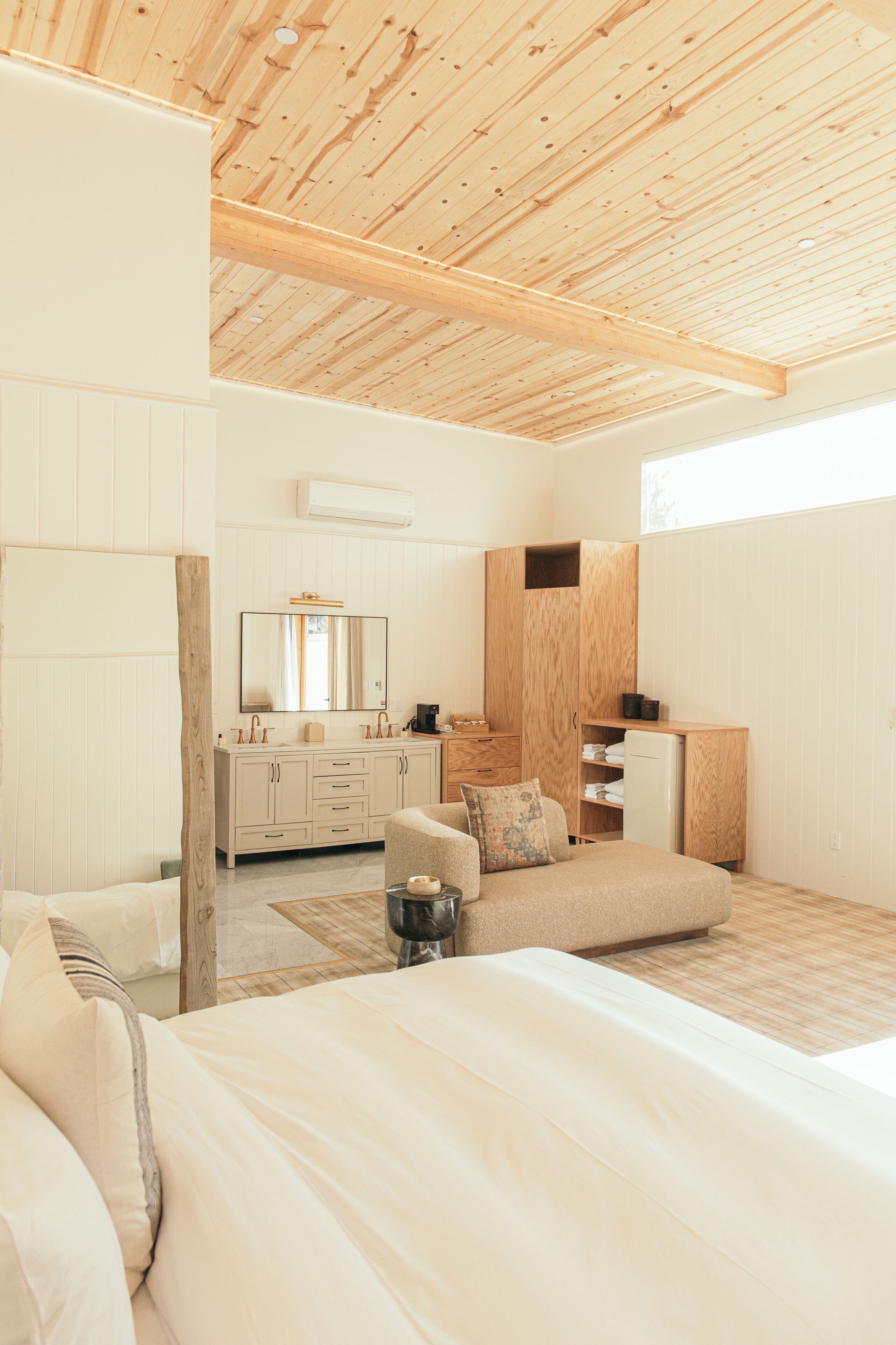 Un dormitorio moderno con techo de madera, una cama grande, un sofá, un aparador y un espejo.