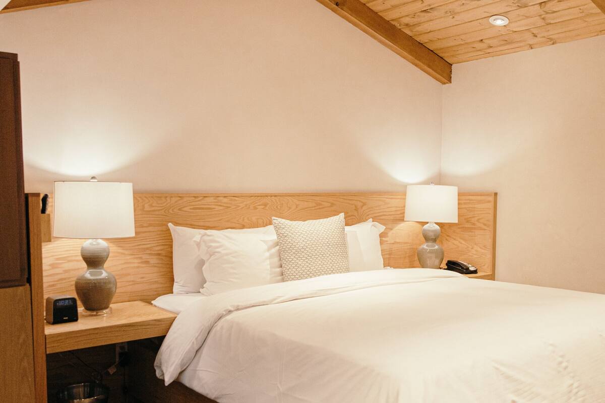 A bedroom with a wooden ceiling, a bed with white bedding, two bedside lamps, and a nightstand with a phone and a trash can.