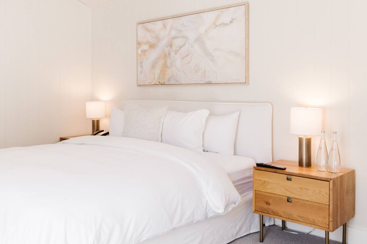 A bedroom with a bed, bedside lamps, a wooden nightstand, a hanging light fixture, and a wall-mounted abstract painting.