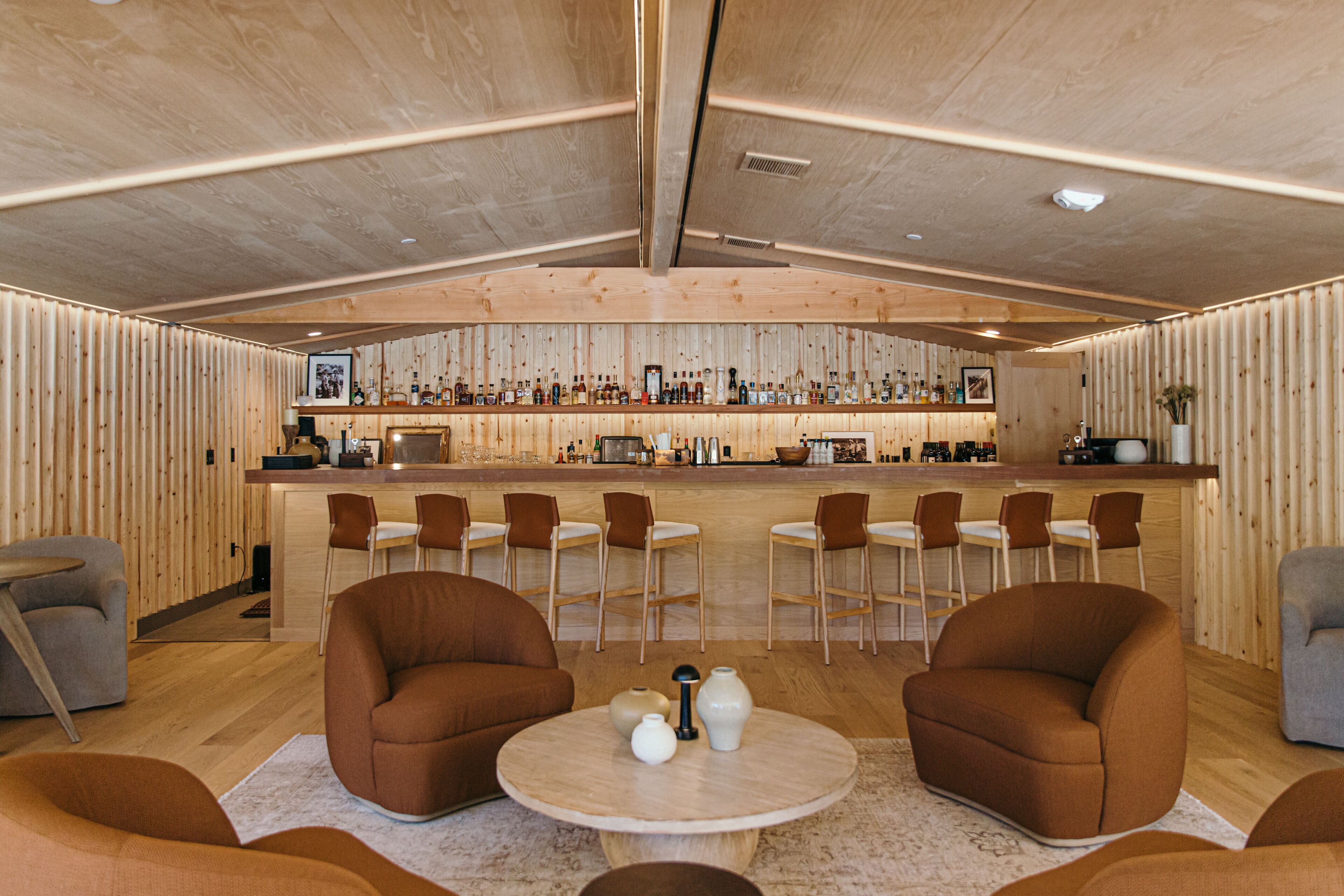 A modern bar area with wooden paneling, bar stools, and a variety of bottles.