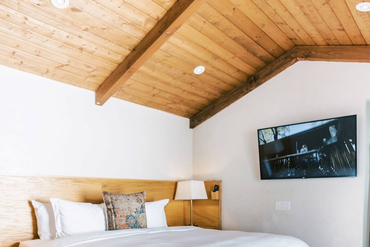 A bedroom with a wooden bed frame, white bedding, a nightstand with a lamp, and a television mounted on the wall.