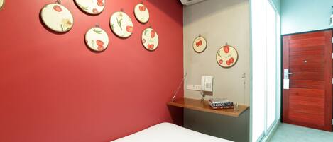 A room with a red wall, a white bed, a wooden shelf with a phone and a book, and a red door.
