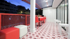 Un'area salotto esterna moderna con mobili bianchi e rossi, un pavimento decorato e vista su edifici sullo sfondo.