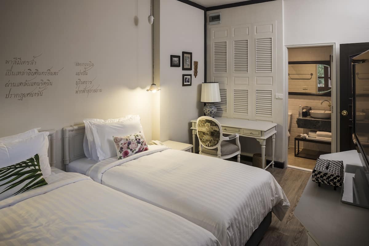 Une chambre d’hôtel avec deux lits, un bureau, une chaise, une lampe et une salle de bain visible à travers une porte ouverte.