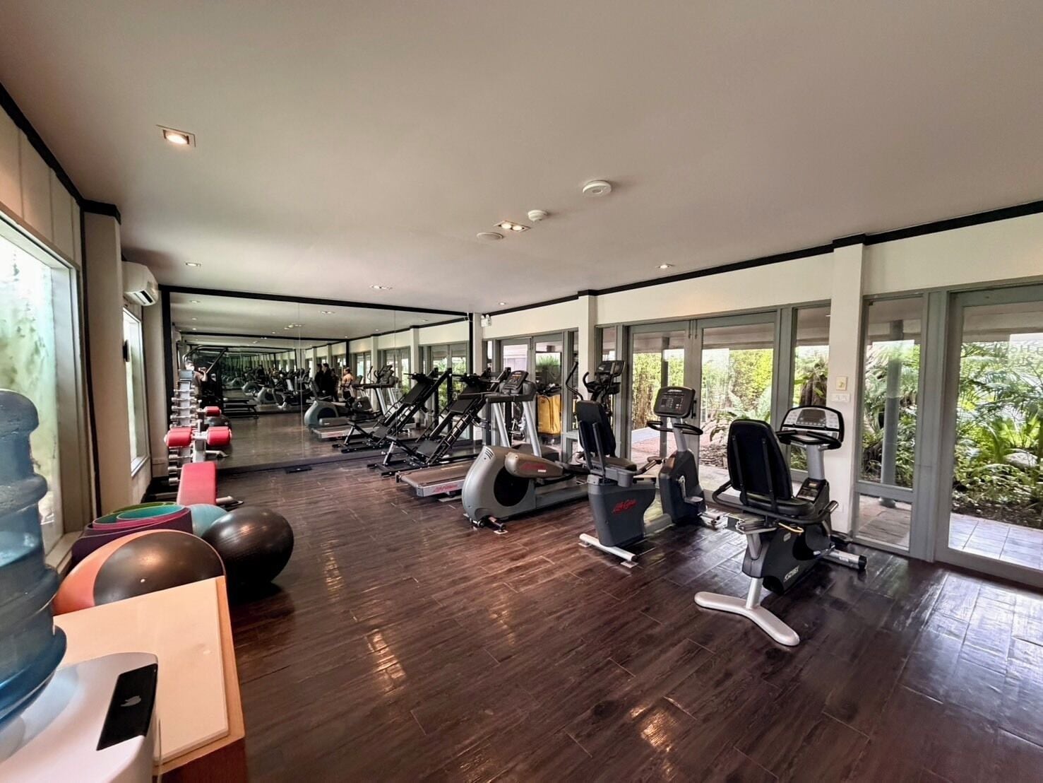 A spacious gym with various exercise equipment, large windows, and a wooden floor.
