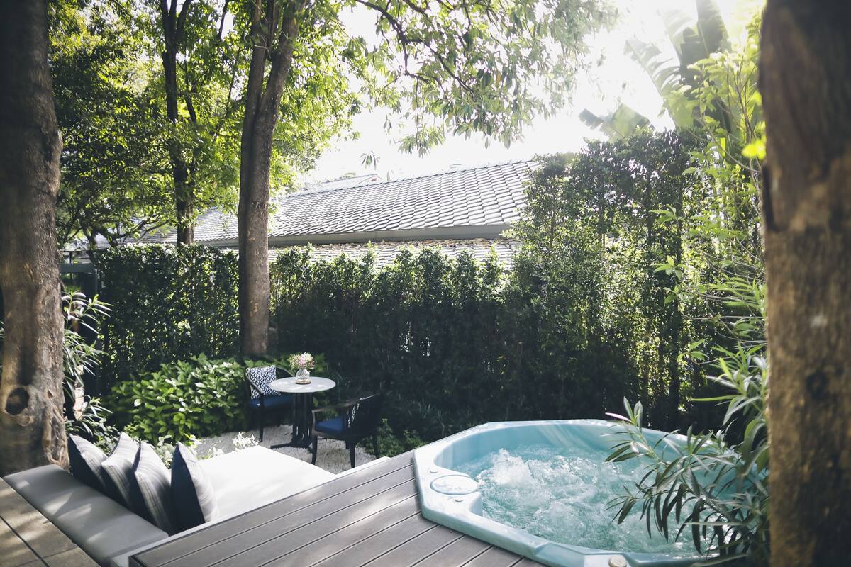 Un bain à remous entouré d’une végétation luxuriante, une terrasse en bois et un coin repas avec des chaises et une table.