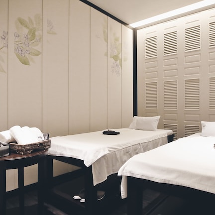 A room with two beds, a small table with towels and a basket, and a wall with floral patterns.