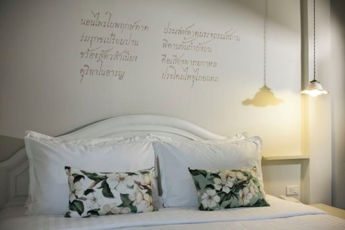 A neatly made bed with white linens and floral pillows, a white headboard, and a wall with Thai script.