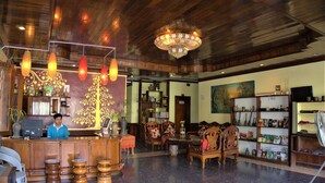 A lobby with a wooden reception desk, multiple chairs, a chandelier, and a bookshelf. - Visoun Namsok Hotel (Luang Prabang)