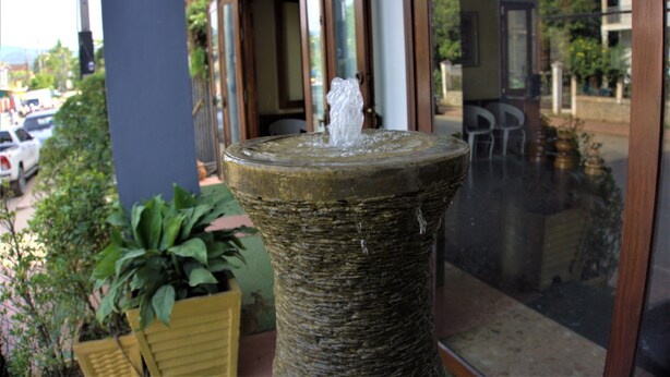 A stone fountain with water spouting from the top, situated on a balcony with a view of parked cars and greenery in the background. - Visoun Namsok Hotel (Luang Prabang)