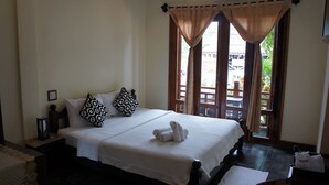 A neatly made bed with white linens and patterned pillows, a bedside table with a lamp, and a window with curtains. - Visoun Namsok Hotel (Luang Prabang)