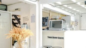A hostel interior with a wooden table, a vase with dried plants, a microwave, and a sign reading "Three Howw Hostel". - Bangkok Nomad Hostel (Bangkok)
