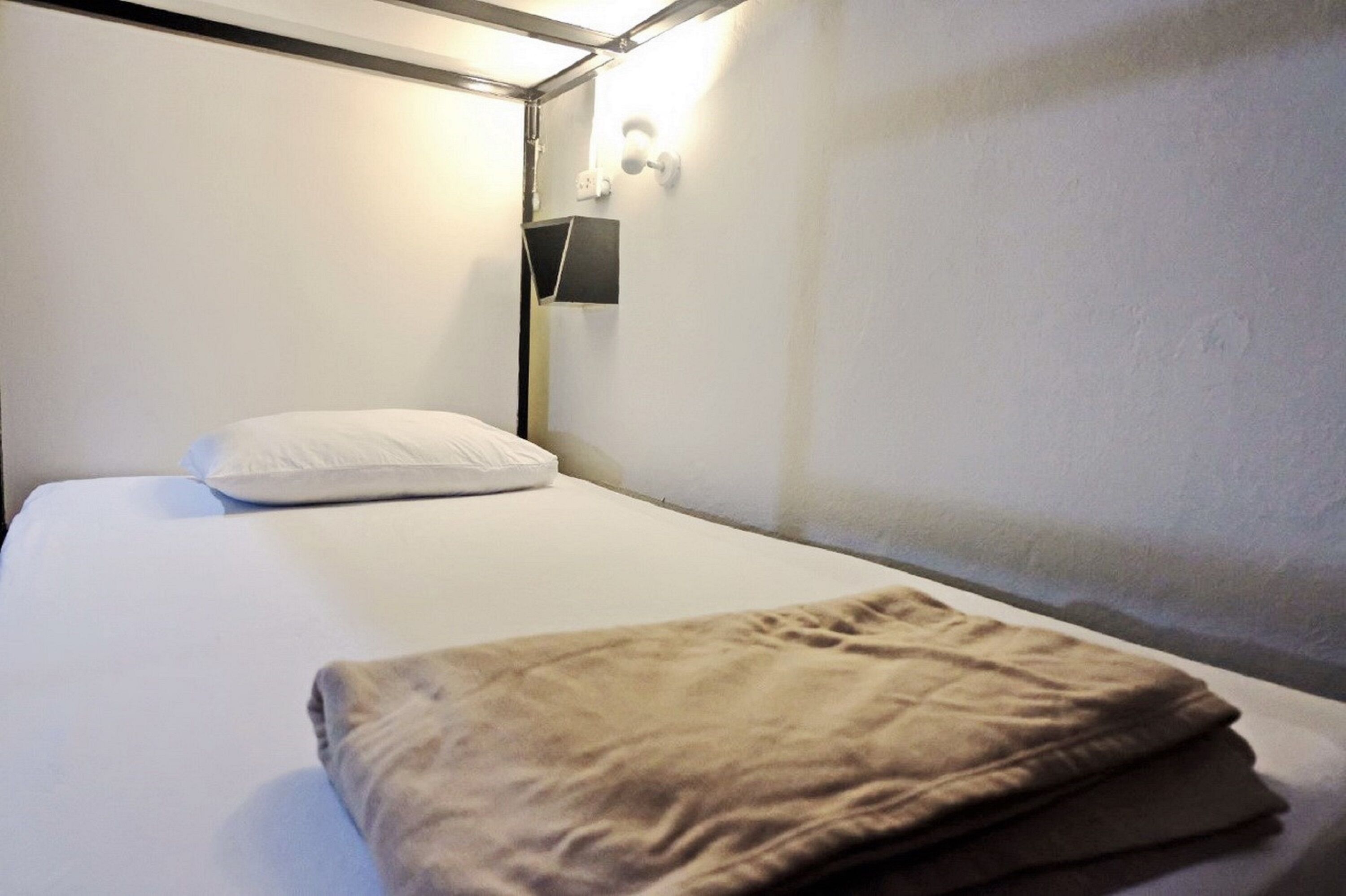 Una cama individual con un colchón blanco y una manta marrón doblada. Hay una lámpara de pared al lado izquierdo de la cama.