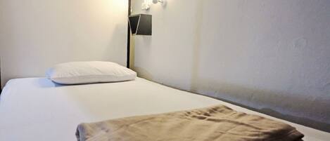 Una cama individual con un colchón blanco y una manta marrón doblada. Hay una lámpara de pared al lado izquierdo de la cama.
