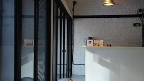 A room with a white brick wall, a black-framed door, a white reception desk, and a clock displaying 16:39. - Bangkok Nomad Hostel (Bangkok)