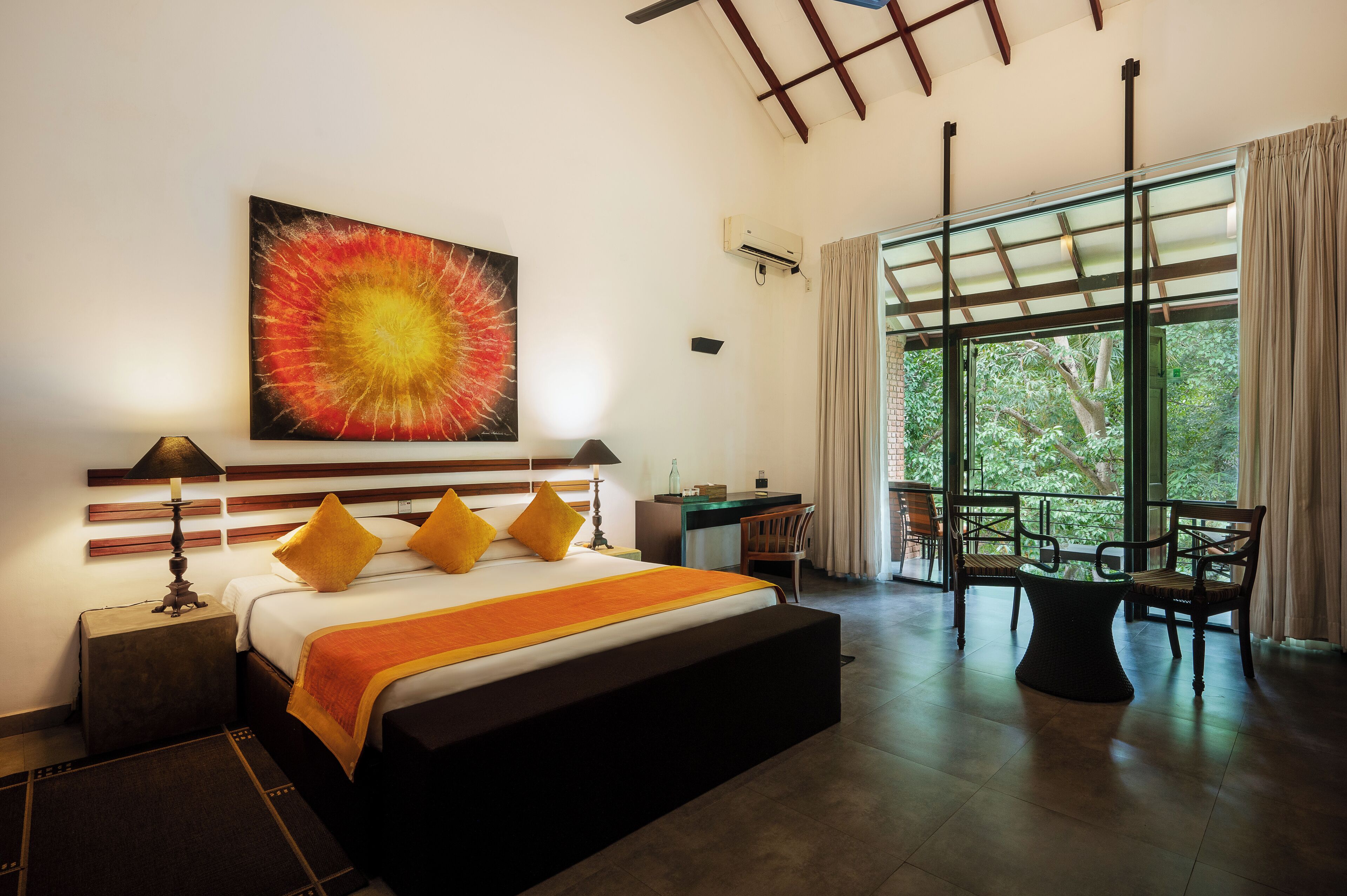 A modern hotel room with a large bed, a desk, and a view of greenery through large windows.