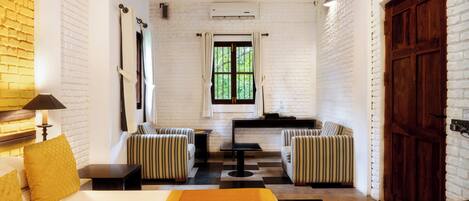 A hotel room with a bed, a small table, two striped armchairs, a window with curtains, a radiator, and a wooden door.