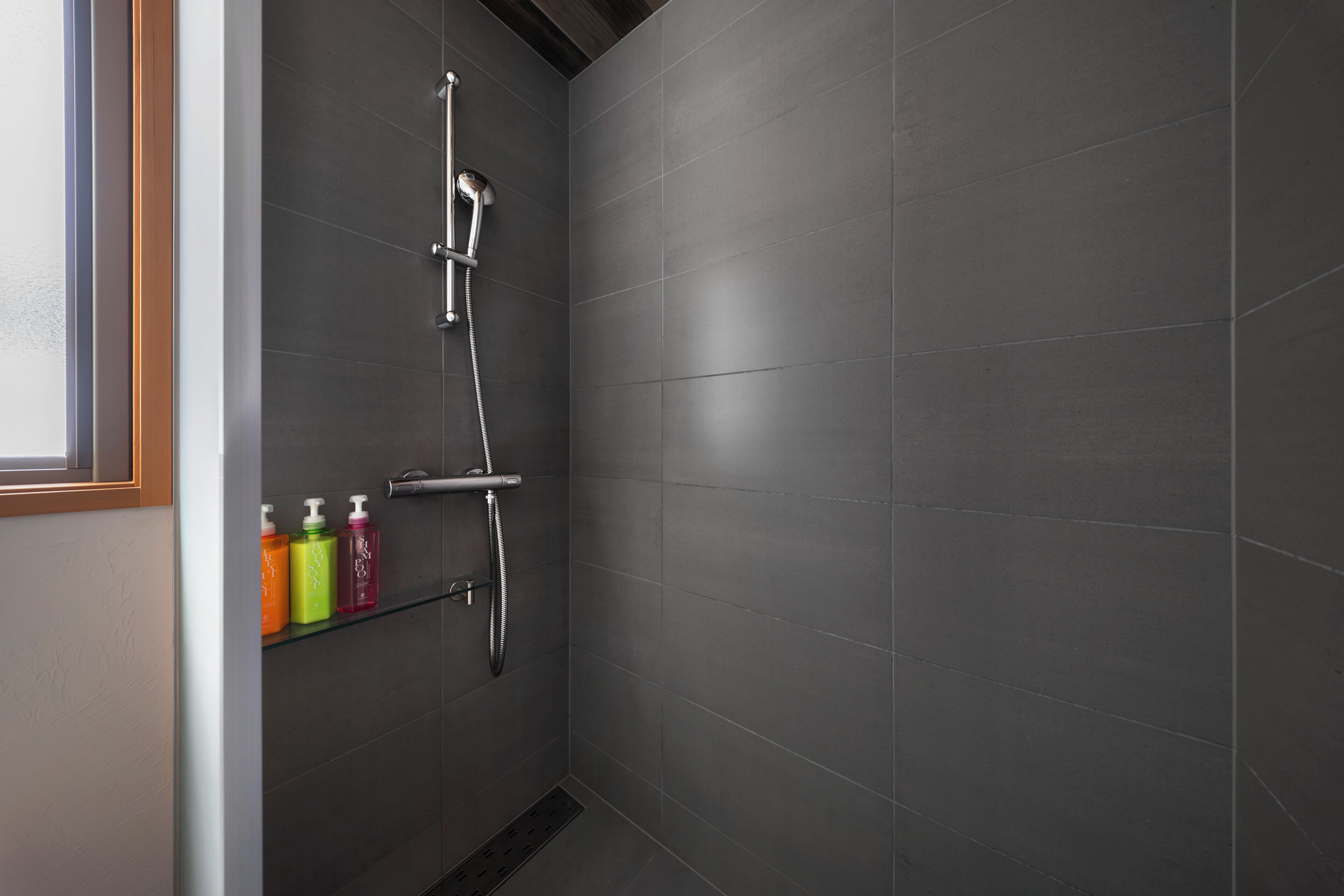 A modern bathroom with dark tiles, a shower with a handheld showerhead, and a shelf with colorful bottles.
