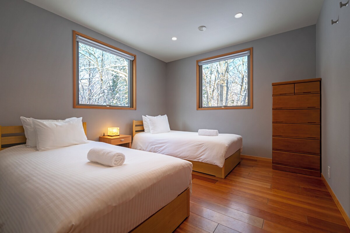 A hotel room with two beds, wooden floors, and large windows with a view of trees.