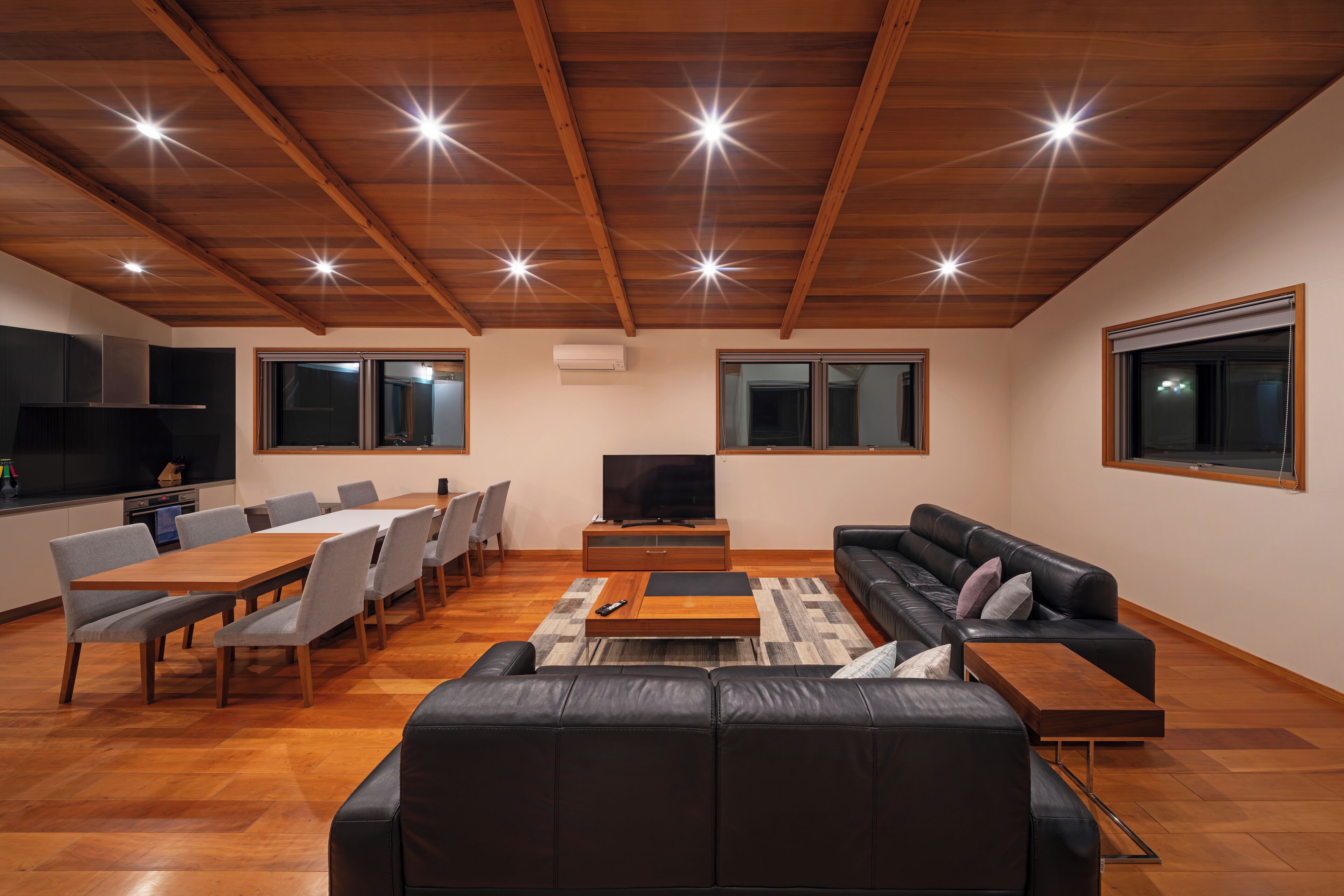 A modern living room with a wooden ceiling, a black leather sofa, a dining table with chairs, and a flat-screen TV.