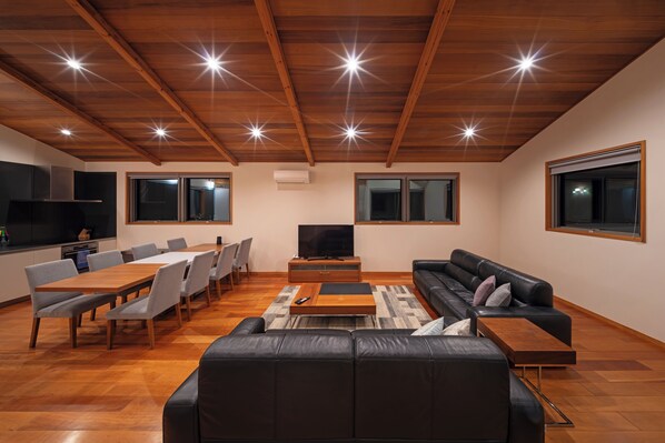 A modern living room with a wooden ceiling, a black leather sofa, a dining table with chairs, and a flat-screen TV. - Bluebird Chalets (Hakuba)
