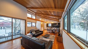 A modern living space with a wooden ceiling, large windows, a black leather sofa, and a kitchen area. - Bluebird Chalets (Hakuba)