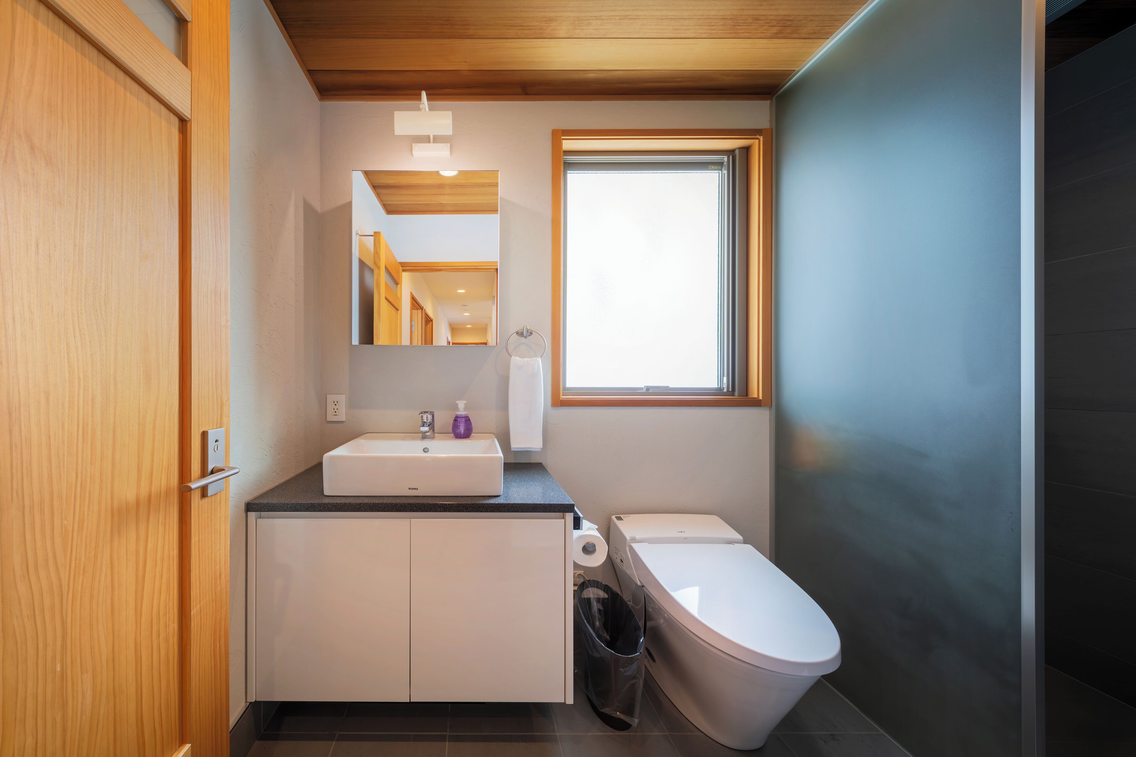 A modern bathroom with a wooden ceiling, a white sink, a toilet, and a window providing natural light.