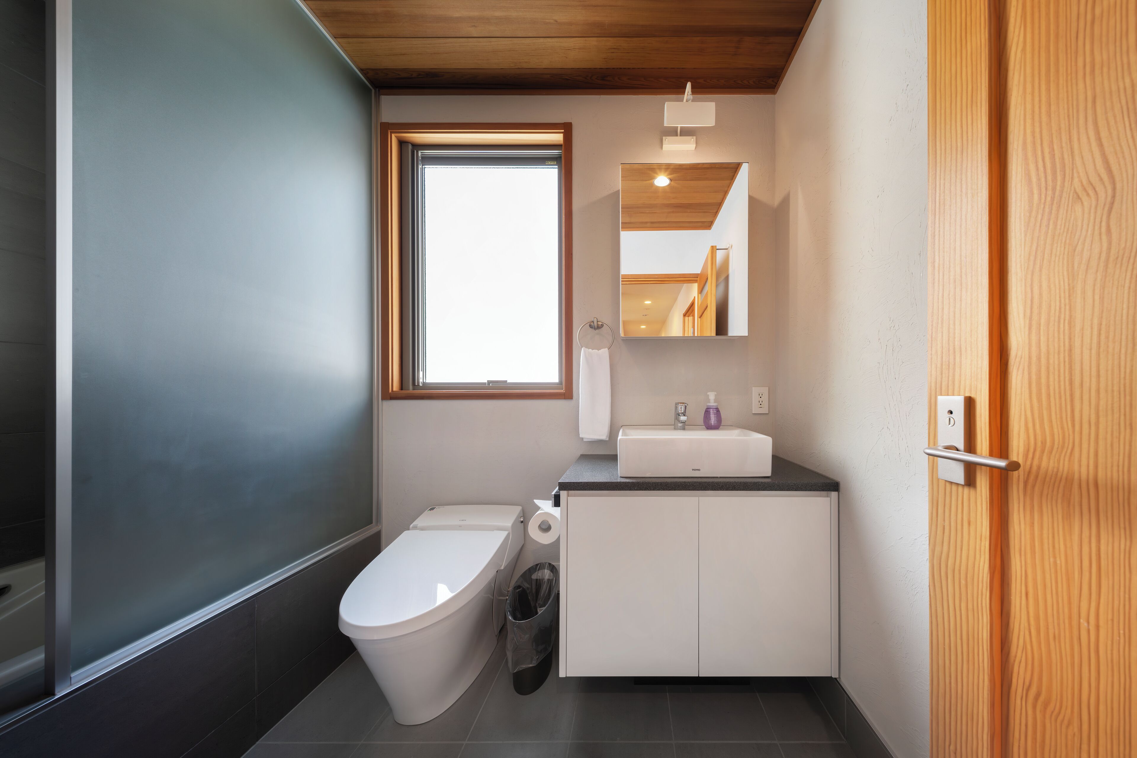 A modern bathroom with a toilet, sink, and mirror. There is a window providing natural light and a door with a handle.