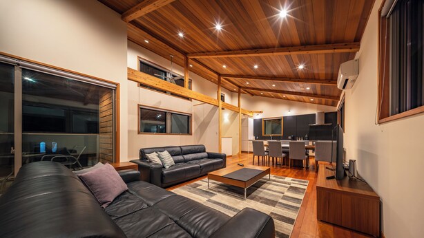 A modern living room with a black leather sofa, a wooden coffee table, and a kitchen area in the background. - Bluebird Chalets (Hakuba)