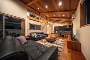 A modern living room with a black leather sofa, a wooden coffee table, and a kitchen area in the background. - Bluebird Chalets (Hakuba)