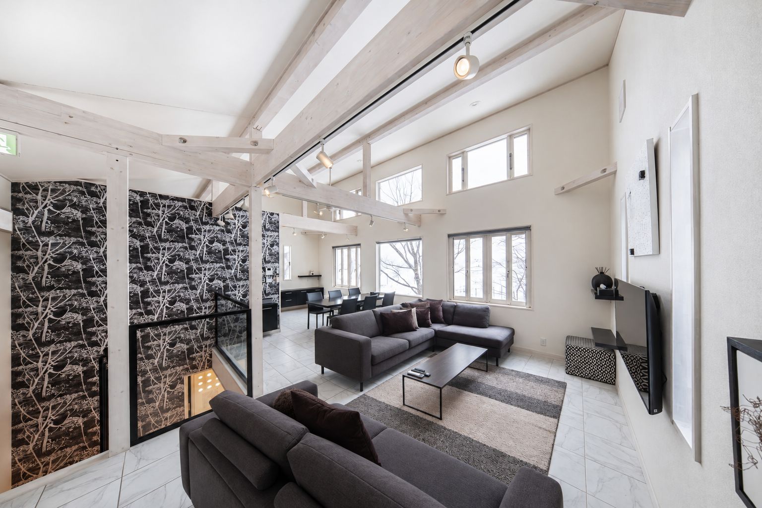 A spacious living area with a large sofa, a coffee table, and a TV mounted on the wall. There are multiple windows providing natural light and a view of the outside.