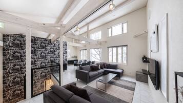 A spacious living area with a large sofa, a coffee table, and a TV mounted on the wall. There are multiple windows providing natural light and a view of the outside.