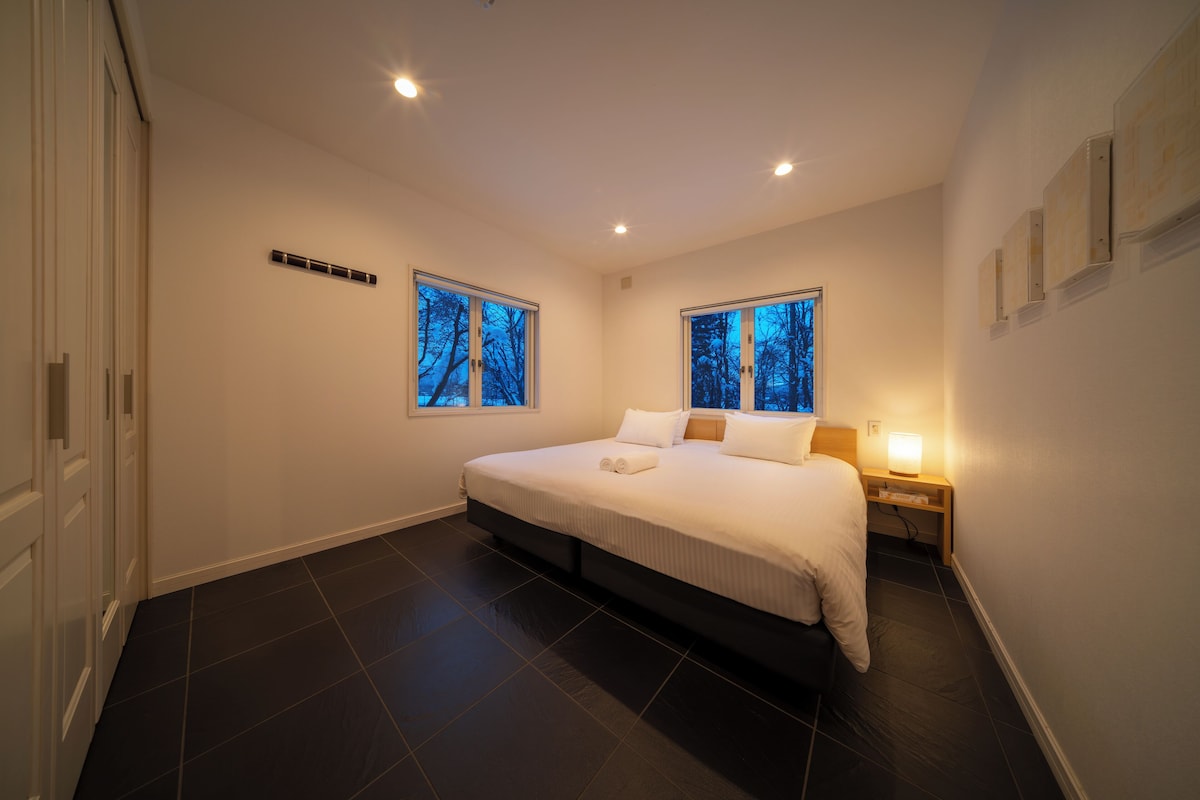 A modern bedroom with a large bed, two windows with a view of trees, a bedside table with a lamp, and a dark tiled floor.
