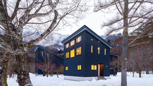 Een modern huis met twee verdiepingen, met een donkere buitenkant, omgeven door een met sneeuw bedekte grond en kale bomen, met op de achtergrond bergen.