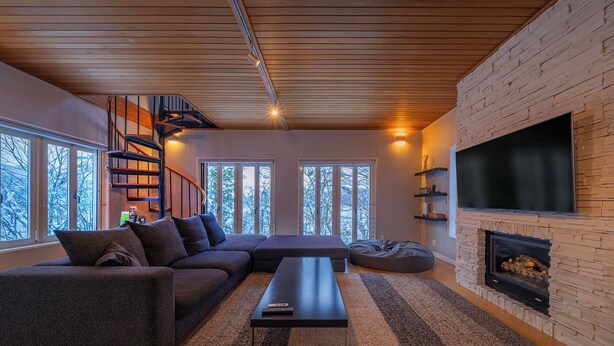 Een moderne woonkamer met een houten plafond, een open haard, een grote televisie en een gezellige zithoek met een bank en een lange zitbank.