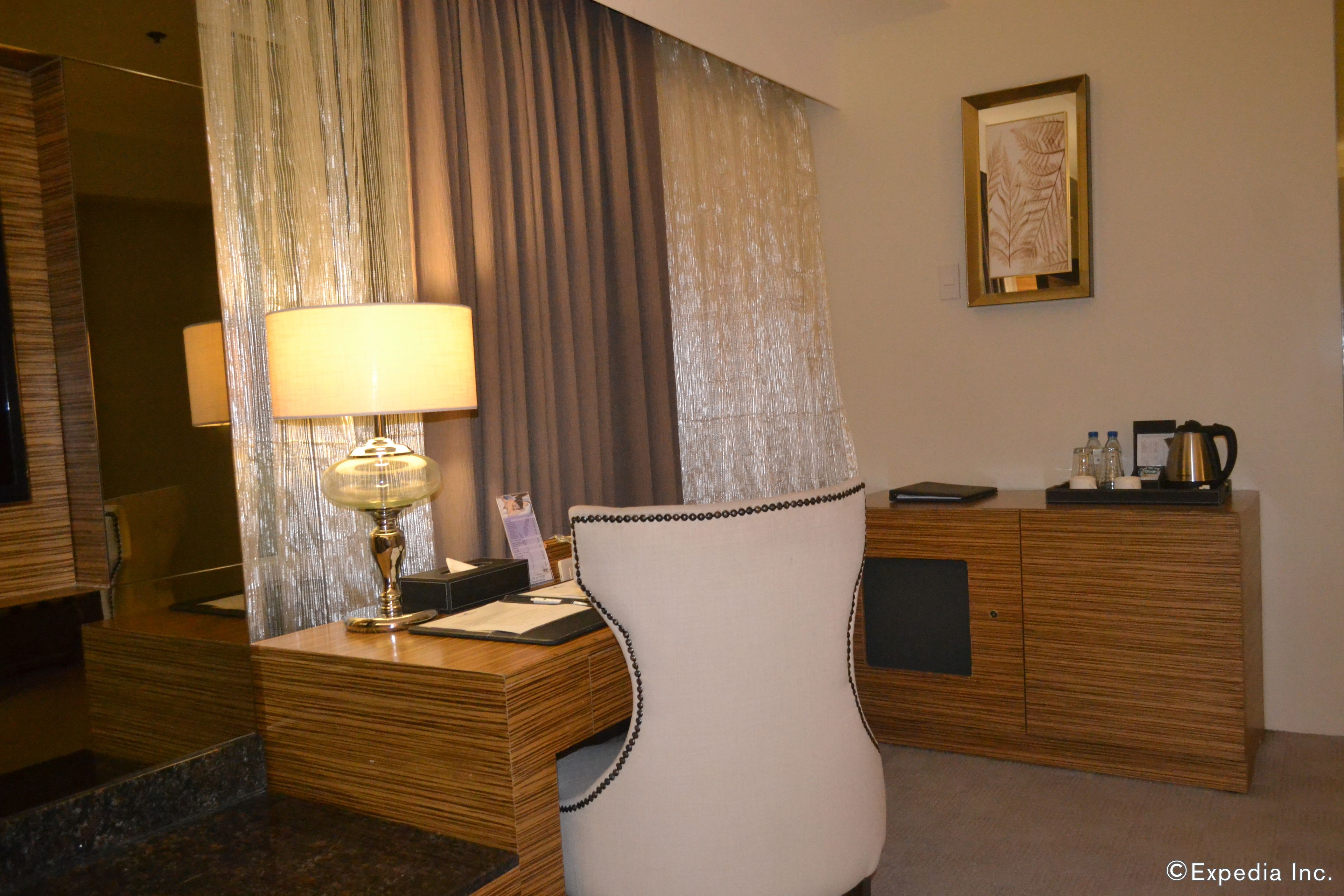 Habitación de hotel con escritorio de madera, una silla blanca, lámpara, minibar y un amplio ventanal con cortinas.
